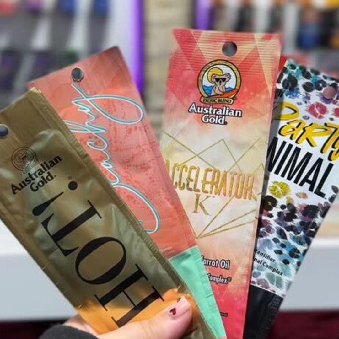Hand holding Australian Gold HOT Accelerator K and Party Animal tanning lotion sachets from The Tanning Shop