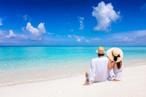 Romantic couple with sun hats sitting on a tropical beach - holiday tan preparation with The Tanning Shop UK