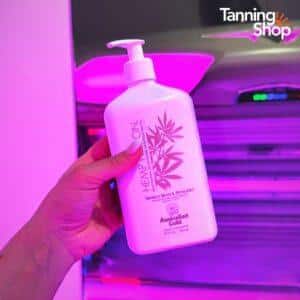 Hand holding Australian Gold Hemp Nation Vanilla Bean and Pistachio moisturising tan extender lotion in front of a pink-lit Tanning Shop sunbed, perfect for extending and maintaining a golden tan.