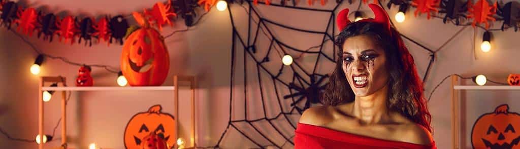 Woman dressed as a devil with red horns and Halloween makeup surrounded by pumpkins and spider web decorations
