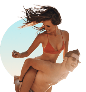 Smiling woman in an orange bikini with her hair blowing in the wind being lifted up by a man in swim shorts on a sunny day, used for The Tanning Shop Student Discount page