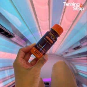 Hand holding a Tanning Boost Vitamin D and Beta Carotene beauty drink inside a Tanning Shop tanning bed lit with blue and pink UV tubes, the ideal tanning supplement for a deeper tan.