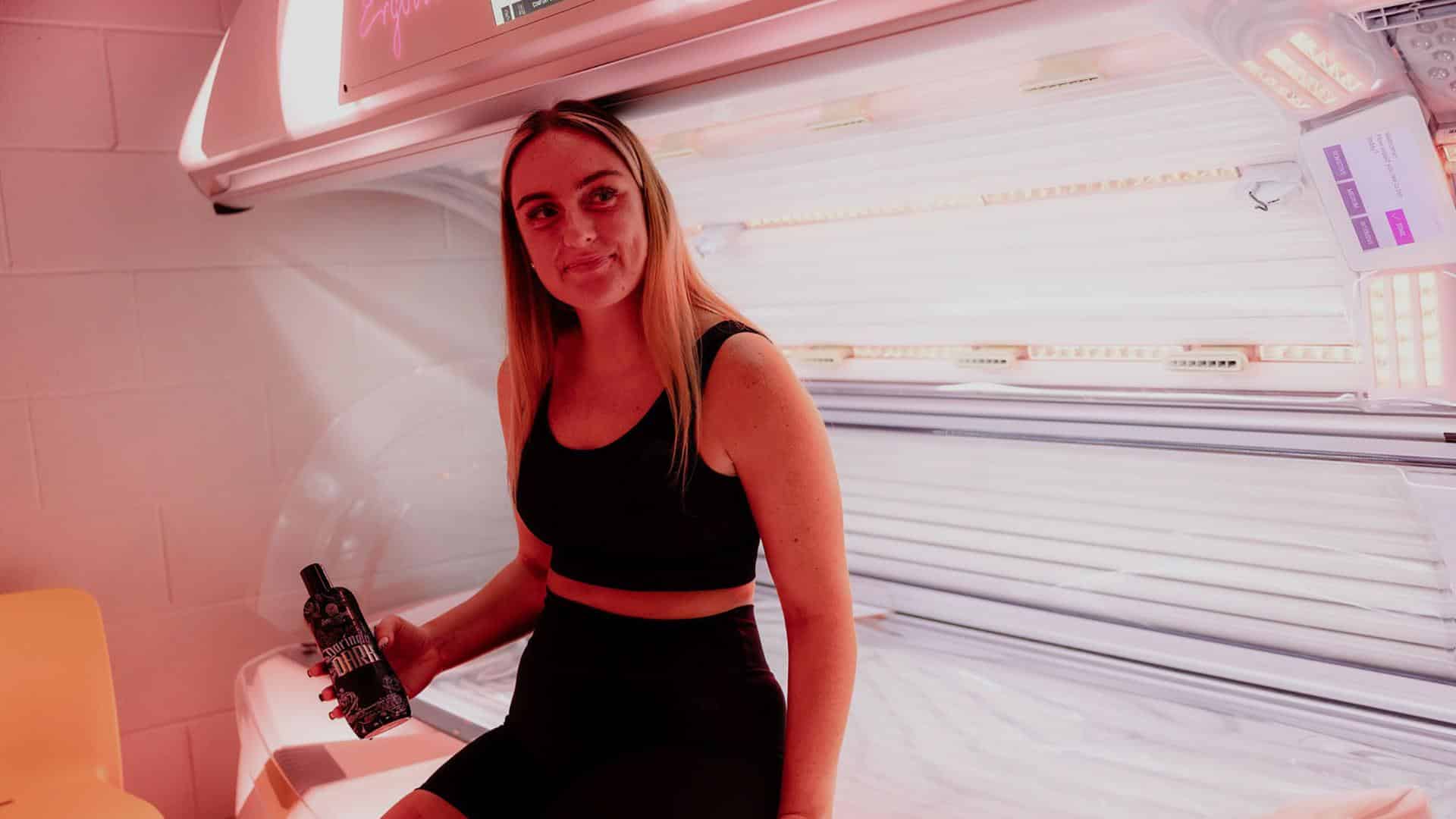 Woman in black activewear sitting next to an open UV tanning bed and holding a tanning lotion bottle at The Tanning Shop