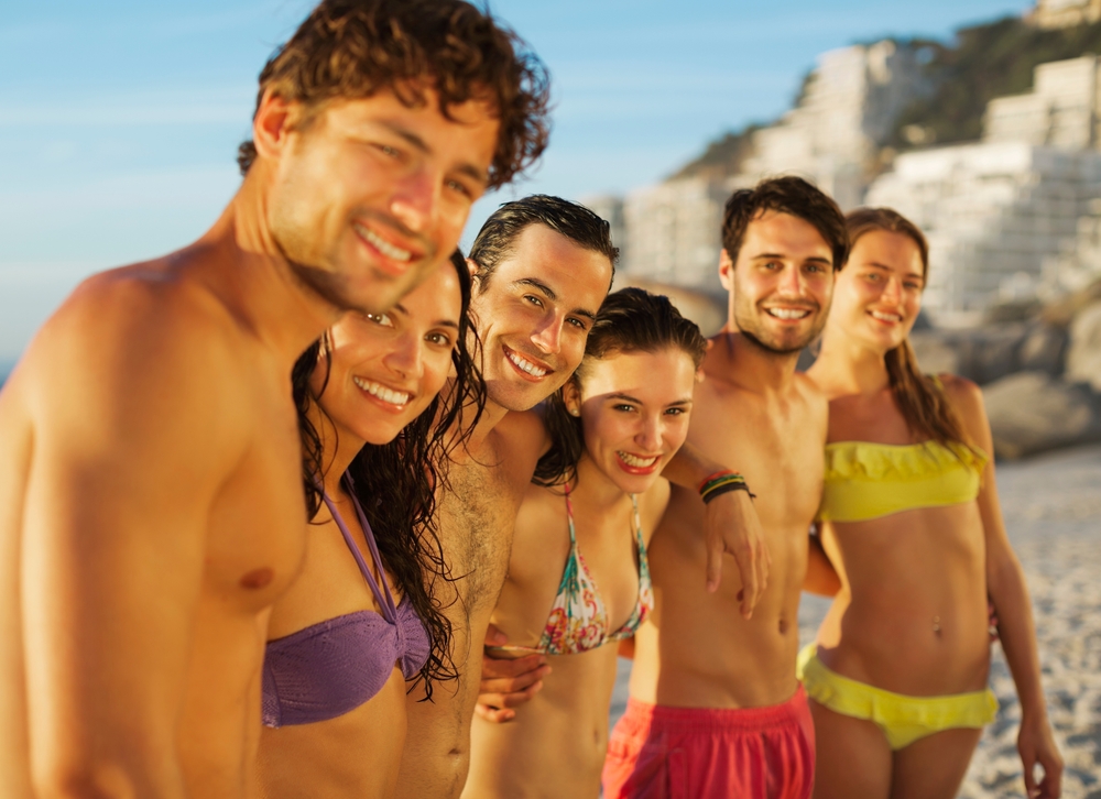 A group of smiling young men and women in swimwear posing together on a sunny beach