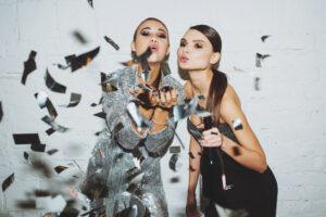 Two women blow silver confetti toward the camera at a glamorous party, one wearing a silver sequin outfit and the other a black dress while holding a wine bottle.