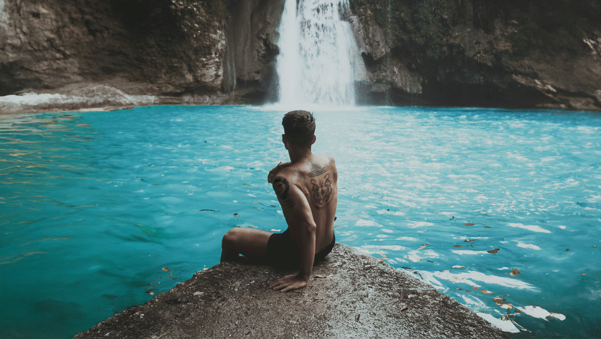 Tanning Shop Influencer Program Tanned Man Waterfall Tanned man sitting by a turquoise waterfall pool, representing the lifestyle and content creation opportunities in The Tanning Shop UK influencer programme